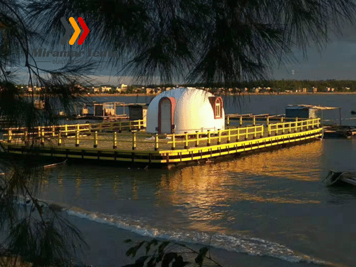 A small Bubbletent on a wooden pier overlooking a body of water at sunset, framed by tree branches in the foreground.