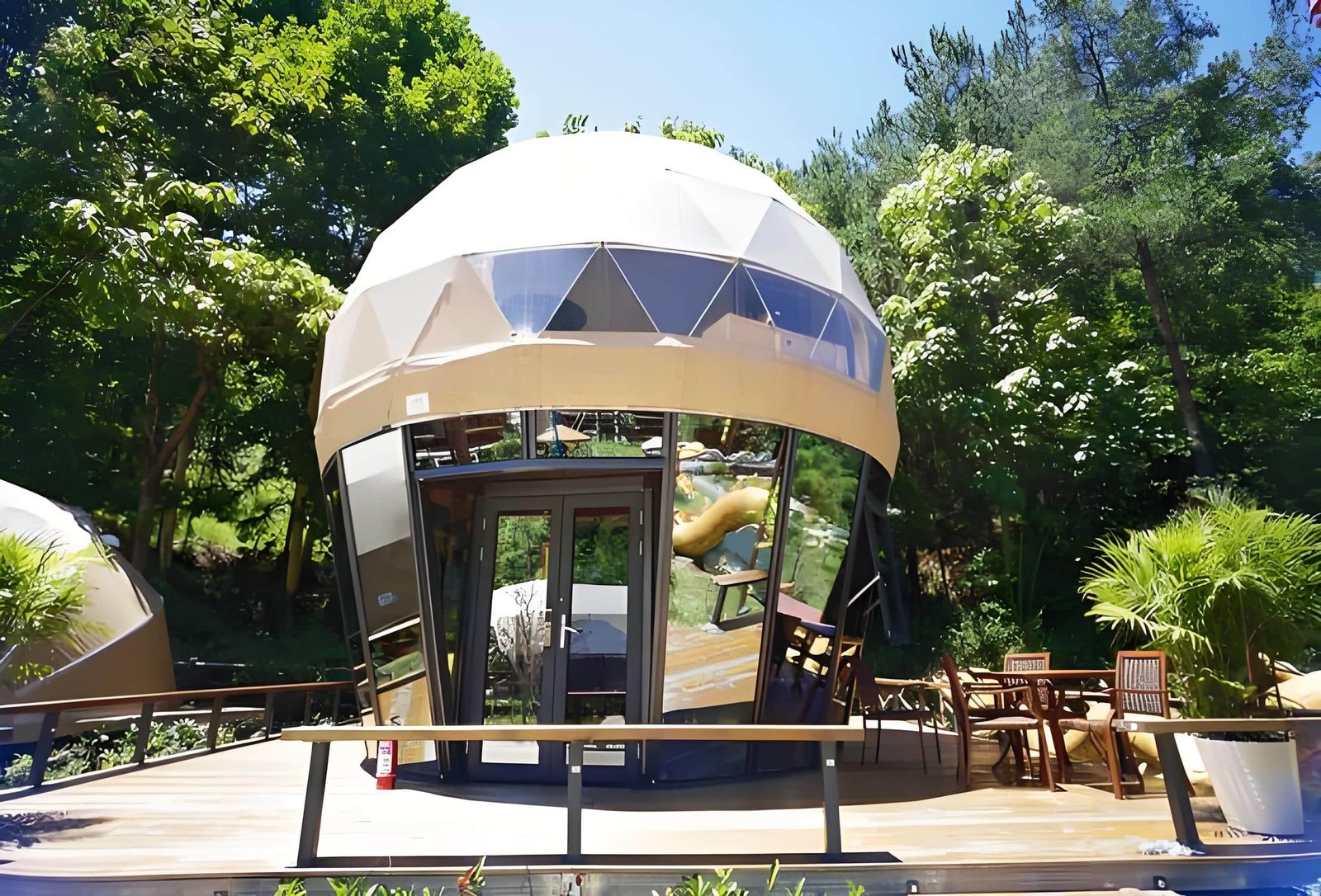 A modern geodesic dome structure with large windows stands amidst lush greenery, featuring a wooden deck with outdoor seating.