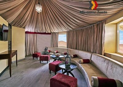Luxurious desert tent interior furnished with plush sofas, an elegant glass table set with drinks, draped ceilings, and panoramic windows with expansive desert views.