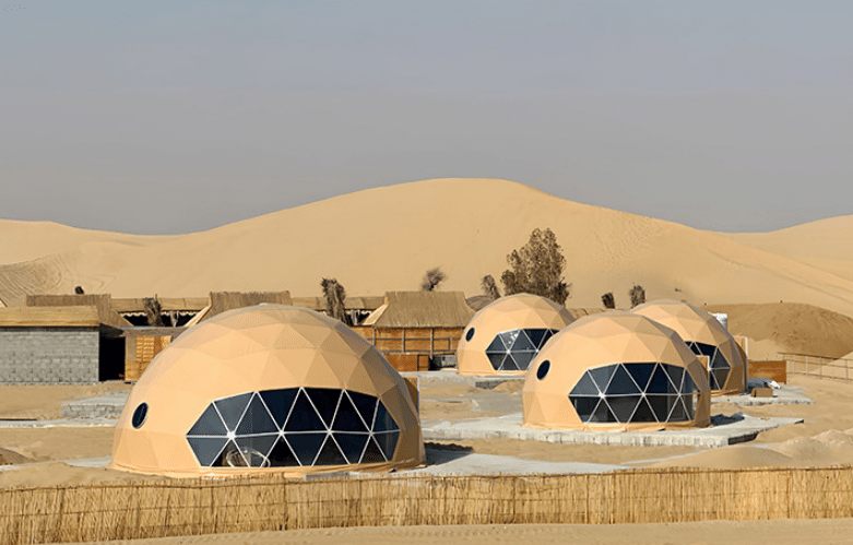 A row of Glamping Dome Tents in a desert setting, perfect for glamping, with sand dunes in the background and sparse vegetation visible.