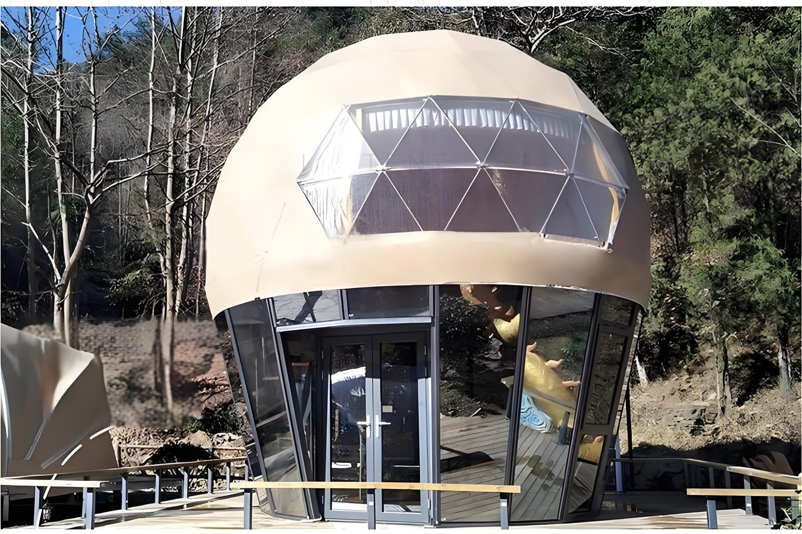 A dome-shaped structure with glass windows stands in a wooded area. It has a geodesic upper part and an entrance with double doors.