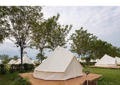 Bell tents on wooden platforms in a grassy area with trees stretch under a partly cloudy sky. A wheeled cart rests in the foreground, adding rustic charm.