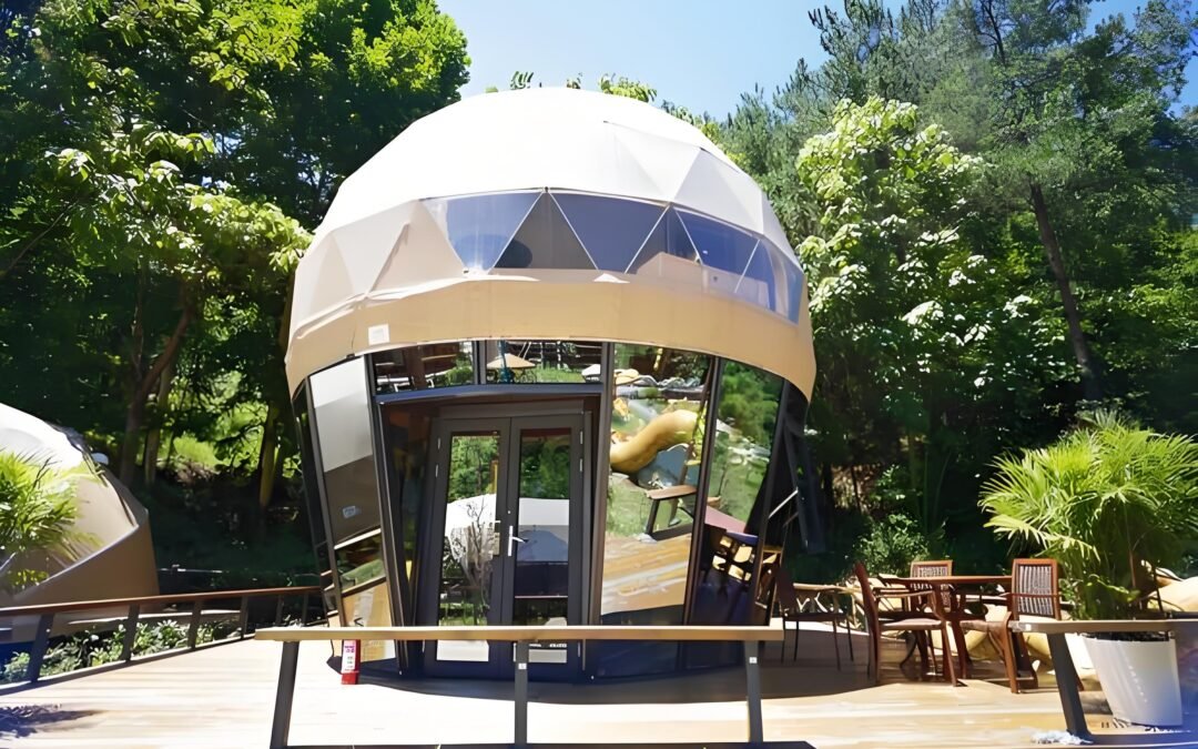 A modern geodesic dome structure with large windows stands amidst lush greenery, featuring a wooden deck with outdoor seating.