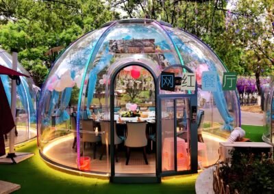 A transparent dining dome with clear walls, elegantly furnished with a table and chairs, is beautifully decorated with balloons and flowers and surrounded by lush greenery.