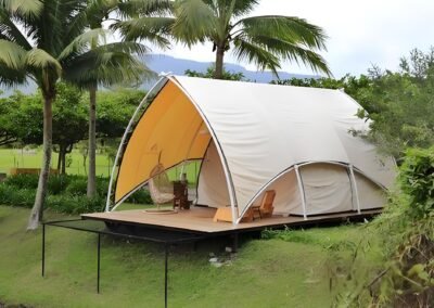 A glamping tent with a curved roof set on a wooden deck, surrounded by palm trees and greenery. It features a hanging chair and a lounge chair inside.