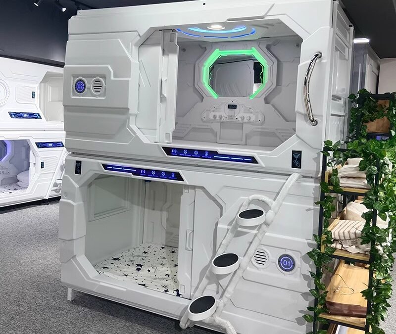 Futuristic Capsule Hotel Room With Illuminated Panels And Ladder Access. Bedding Is Visible Inside, Surrounded By Greenery And Modern Decor.