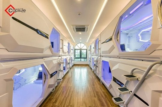 A Hallway Lined With Two Rows Of Futuristic Capsule Beds, Stacked Two Levels High, Featuring Blue Lights And Control Panels, With A Wooden Floor And A Window At The End.